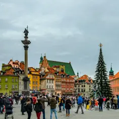 Tłum ludzi na placu w jednym z polskich miast, z kolumną Zygmunta i kolorowymi kamienicami w tle.