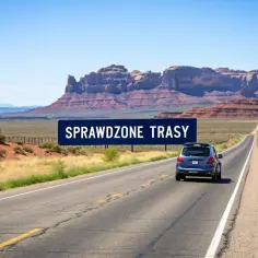 Niebieski samochód jedzie pustą drogą przez krajobraz przypominający Monument Valley, gdzie w oddali widać skalne formacje. Znak "SPRAWDZONE TRASY" sugeruje podróż przez amerykańskie miasta.