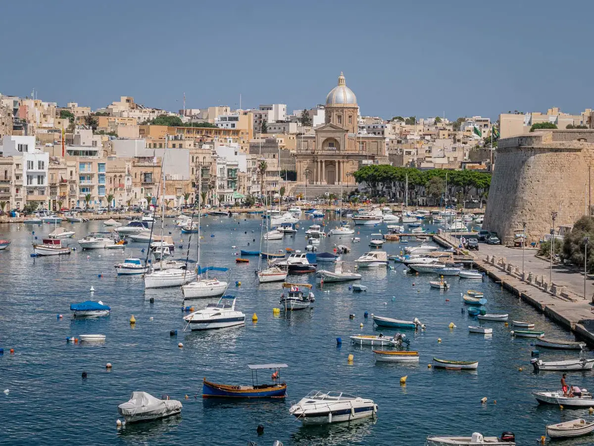 Malta co zobaczyć: malowniczy port pełen łodzi, z charakterystyczną kopułą kościoła i kamienną fortecą w tle.
