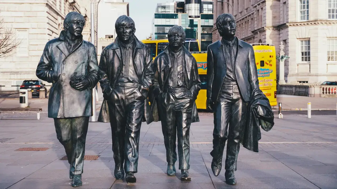 Pomnik Beatlesów w Liverpoolu, mieście beatlesów. Cztery postacie idą chodnikiem, za nimi żółty autobus turystyczny.