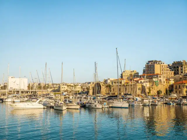 Port w Heraklionie, gdzie cumują jachty, z widokiem na kamienne mury i budynki starego miasta.